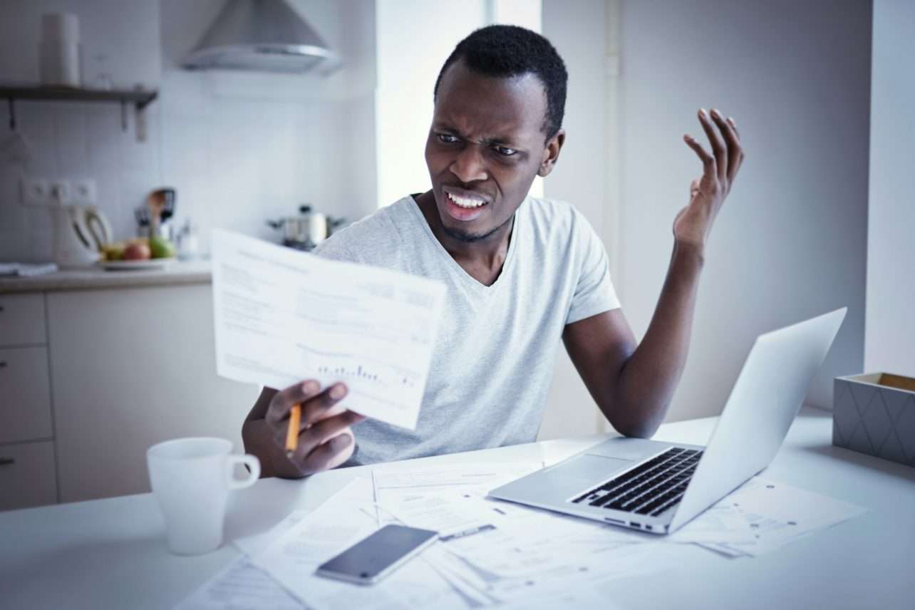 Young african man looking at bill, frustrated about high taxes, having problems with debt